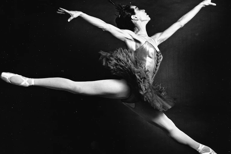 A ballet dancer taking a leap with her toes pointed outward and arms raised.