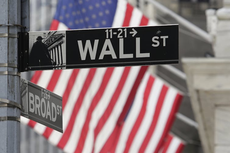 A street sign that says 'Wall St' seen in front of an American flag
