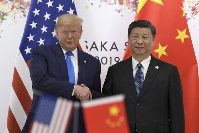 A white man shakes the hand of a Chinese man, each respectively standing in front of an American flag and a China flag
