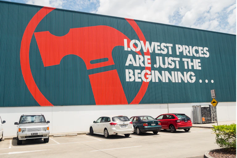 Cars parked in front of building painted with a large red hammer and the slogan: "Lowest prices are just the beginning..."