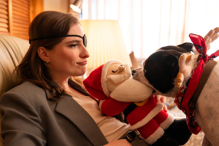 A young women with an eye patch sits on a couch with a Santa plush and a festively dressed dog on her lap.