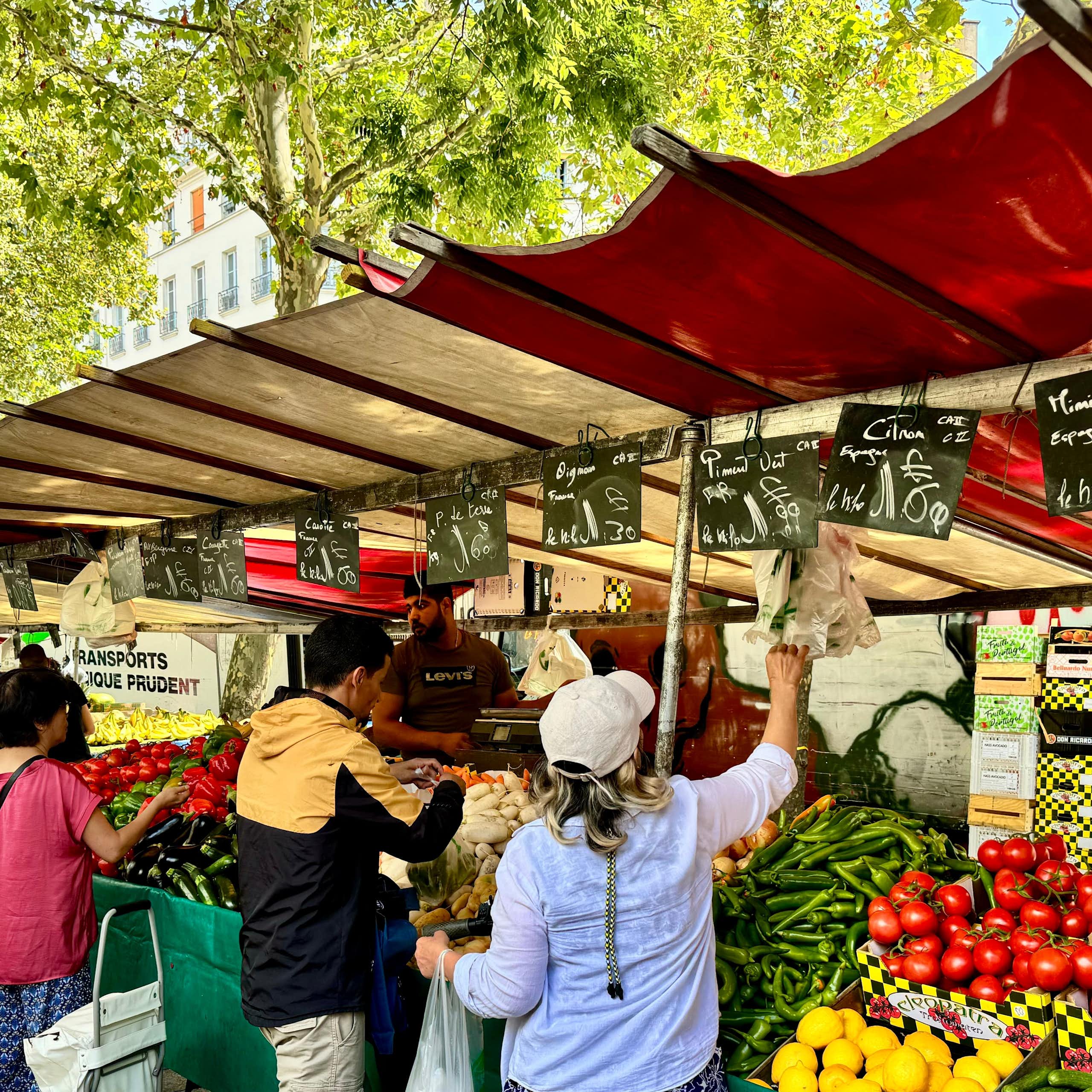 Image d'un marché en plein air avec un étal de fruits et légumes avec des ardoises sur lesquelles sont écrites des prix
