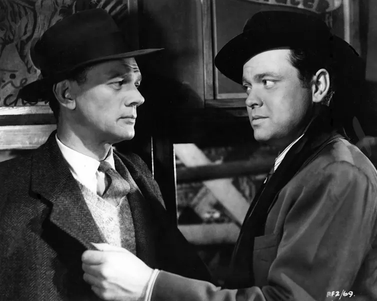 Black and white image of two men dressed in 1940s hats and overcoats.