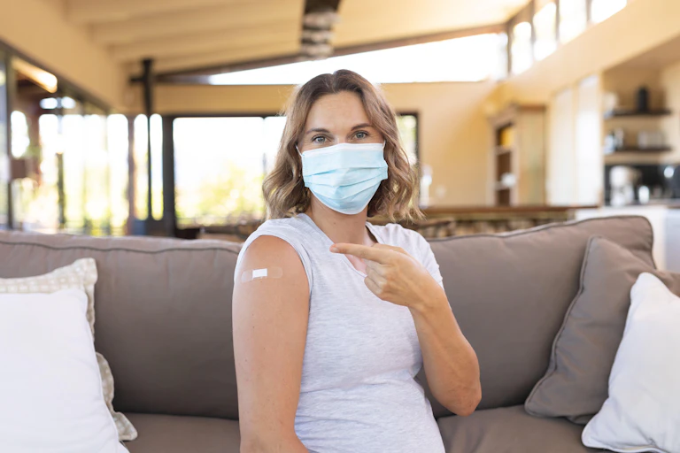 Woman wearing a mask pointing at a plaster on her upper arm from where she had a vaccination.