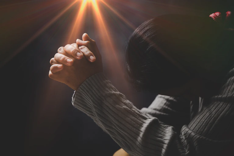 A woman with folded hands against a black background with a yellow light coming from above them
