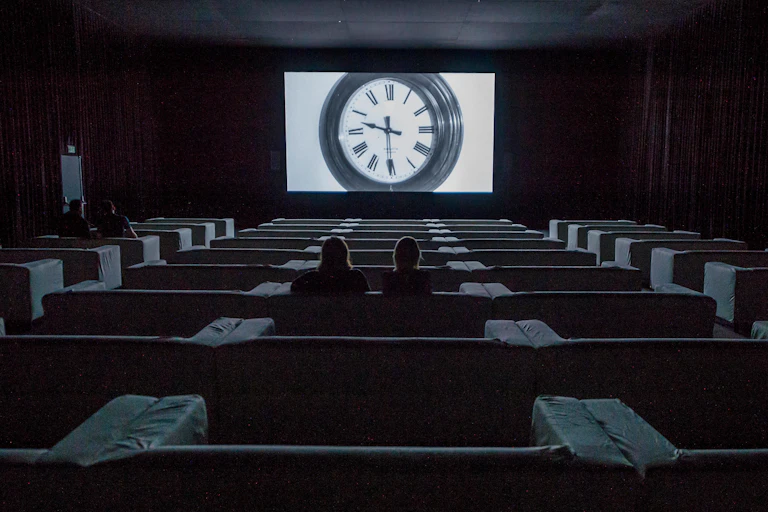 Two people sitting in a cinema with a giant clock on screen.