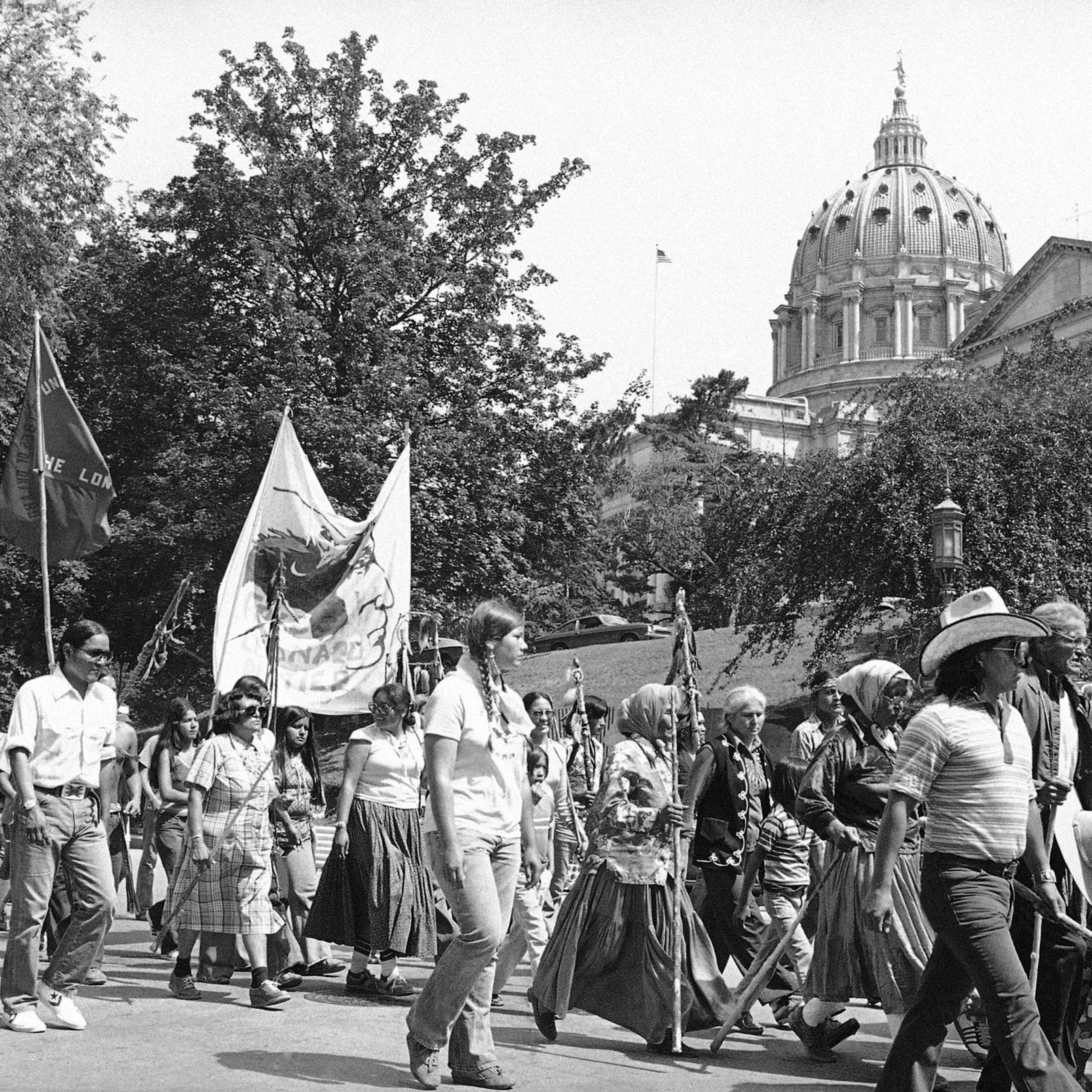 A black and white photo of marchers in warm weather passing a building with a high dome.