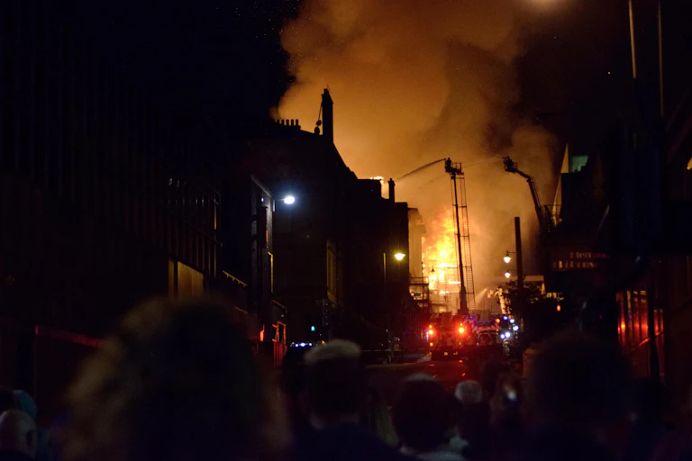 A fire rages over a building with firefighters on ladders trying to put it out, blazing bright orange against the night sky.