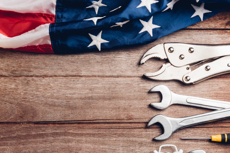 An American flag lies on a wooden table next to a hammer and several wrenches.
