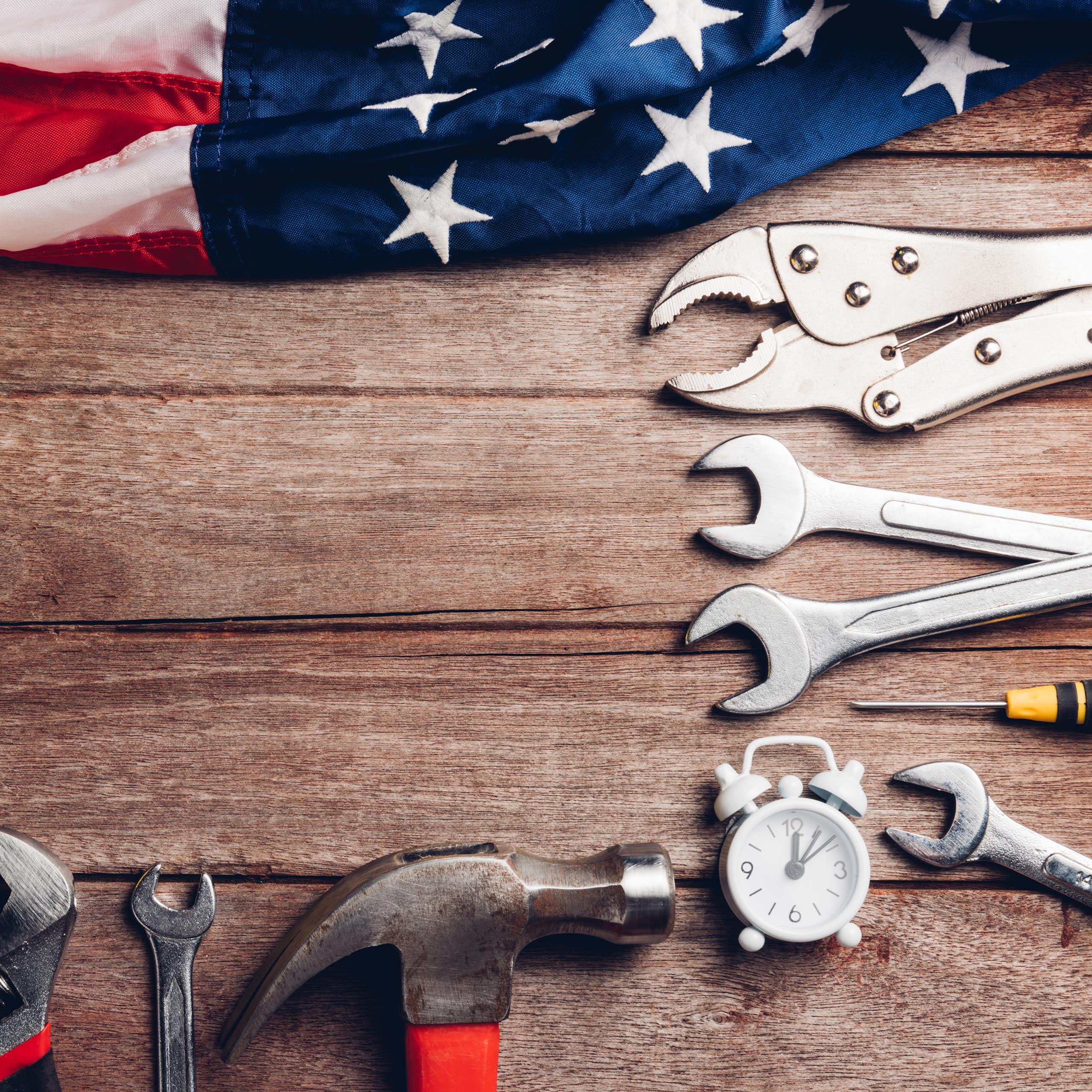 An American flag lies on a wooden table next to a hammer and several wrenches.