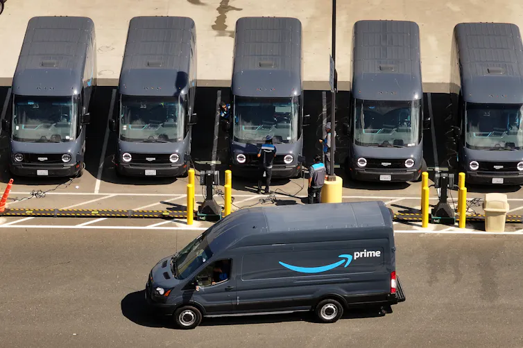 A line of Amazon electric delivery trucks charging while one drives away.
