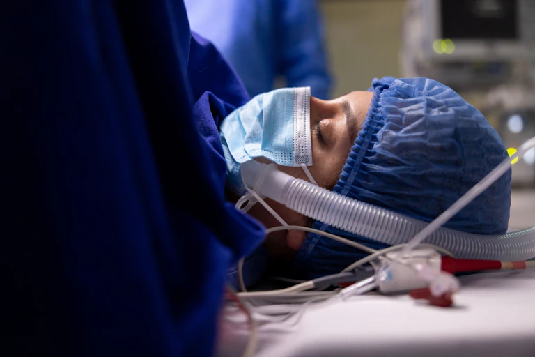 Patient wearing face mask and surgical cap, eyes closed, connected to tubes