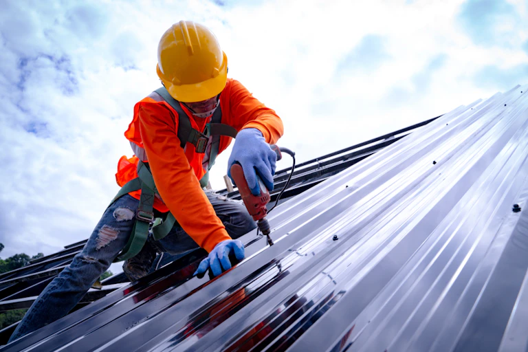 Construction worker on roof, wearing harness and safety gear, fastening roof sheets
