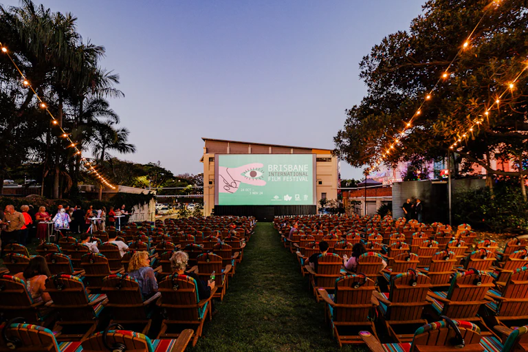Some poeple are seated at an outdoor cinema, qwith fairy lights hanging above and a dimming evening sky.