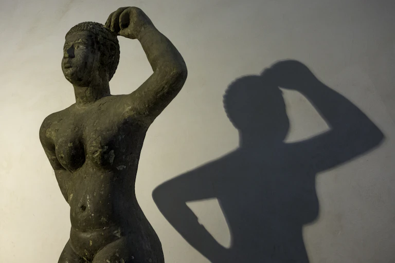 An ancient statue of a woman with an arm raised, her shadow cast on the wall behind her.