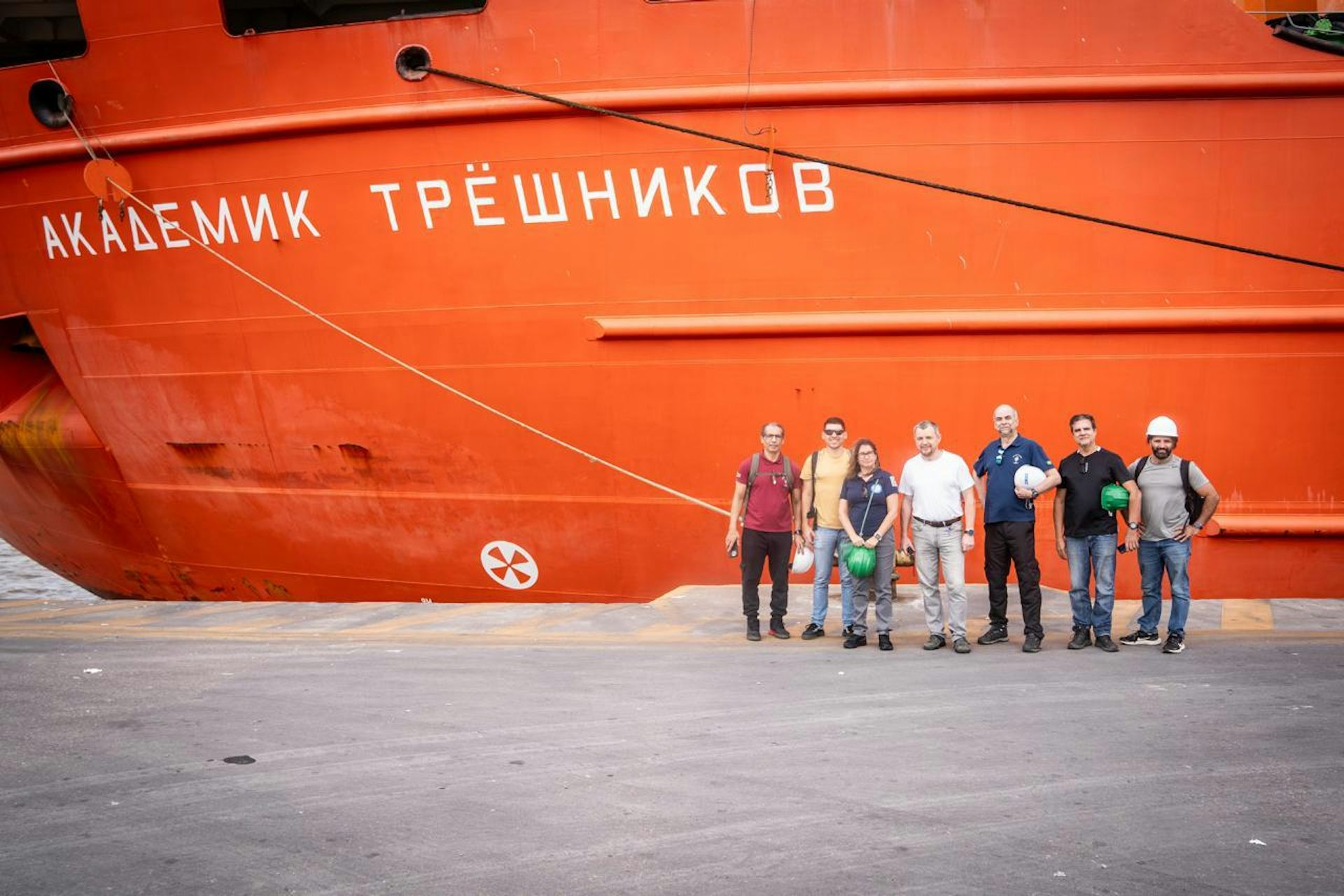 Foto mostra equipe de sete pesquisadores em frente a um navio vermelho, onde se lê, em russo, o nome Akademik Tryoshnikov