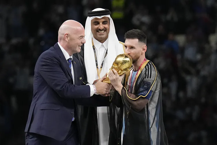 A soccer player kisses a gold trophy as a man in a suit and an emir look on smiling.