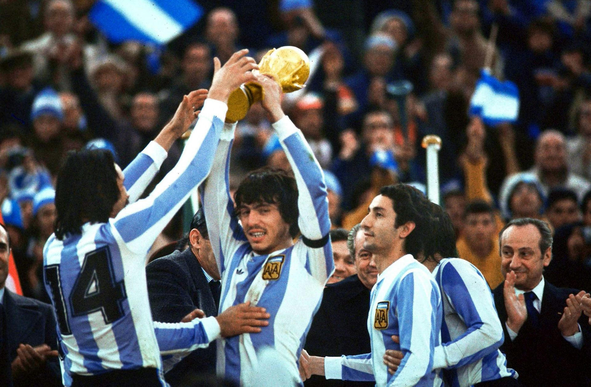 Men in blue-and-white striped soccer jerseys celebrate while holding up a gold trophy.