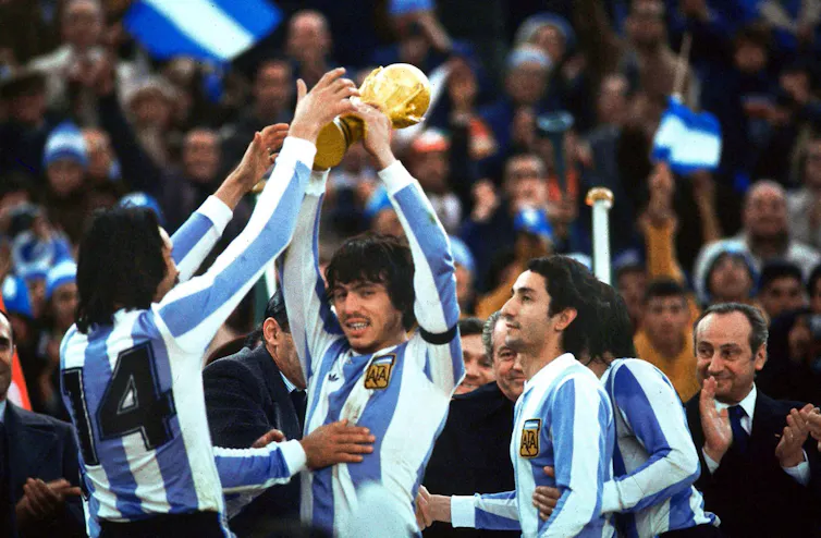 Men in blue-and-white striped soccer jerseys celebrate while holding up a gold trophy.