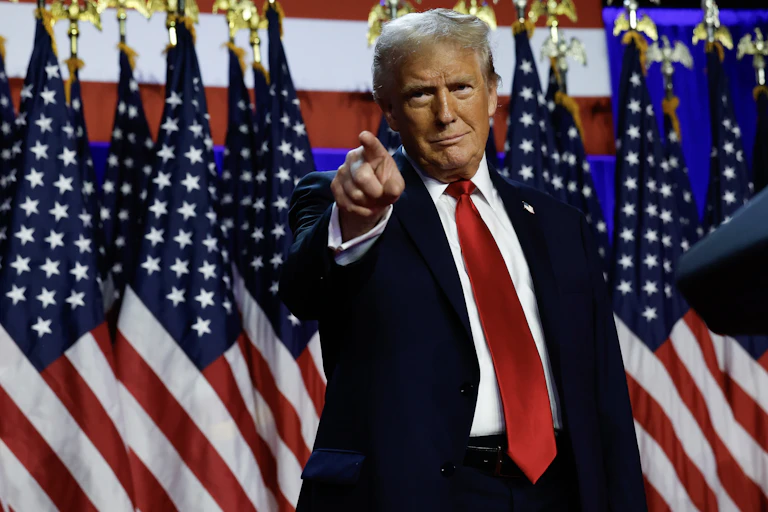 Man in suit and red tie points ahead with his right index finger. Behind him are several American flags.