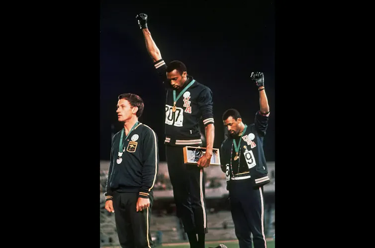Two black men on a medal podium raise their fists and bow their heads.