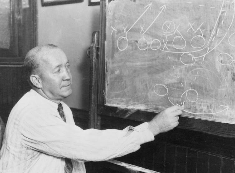 Black and white photo of balding, middle-aged man drawing on a chalkboard.