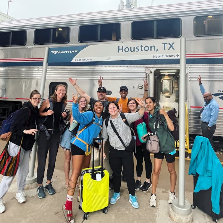 A group of young people smile and pose under a sign that says 'Houston Texas' in front of a train.