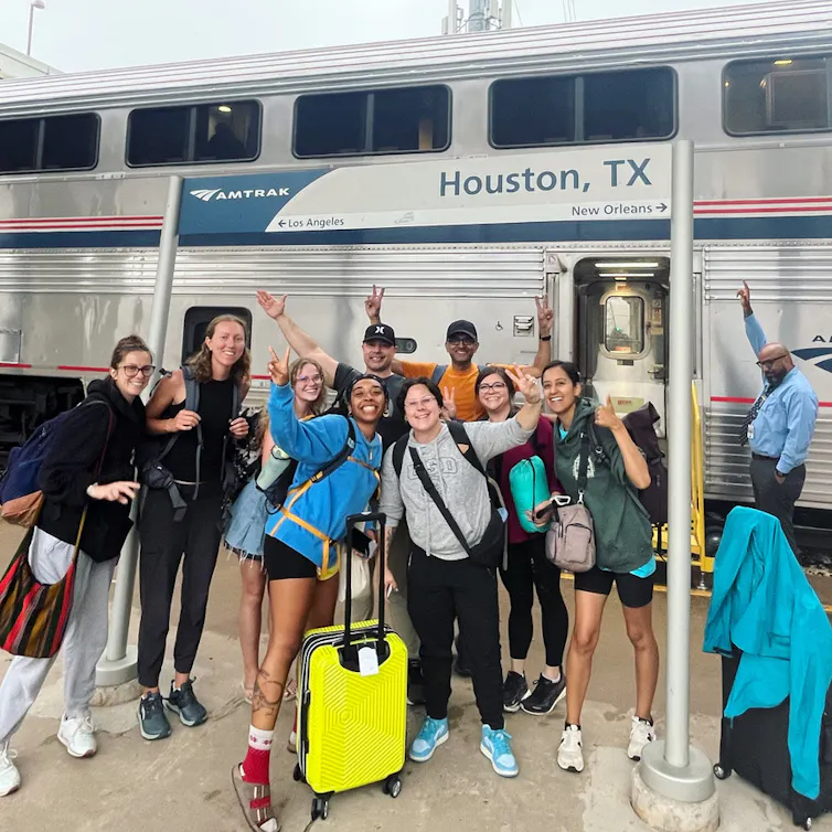 A group of young people smile and pose under a sign that says 'Houston Texas' in front of a train.
