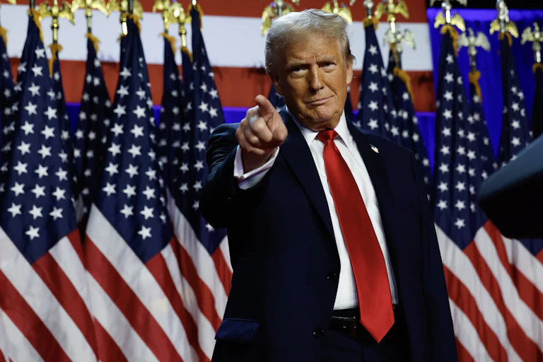 A man in a blue jacket, white shirt and red tie, standing in front of many American flags and pointing away from him.