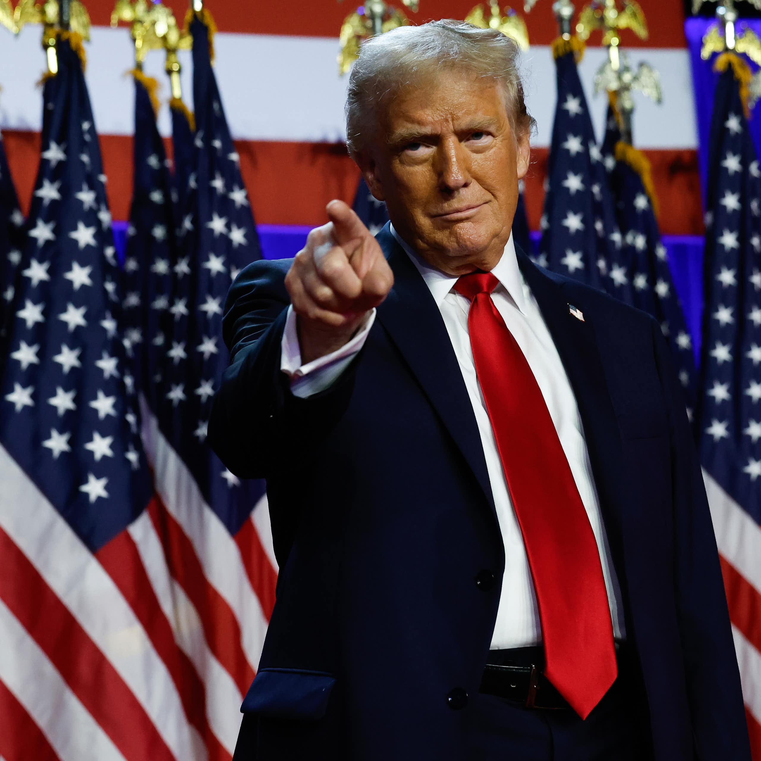 A man in a blue jacket, white shirt and red tie, standing in front of many American flags and pointing away from him.