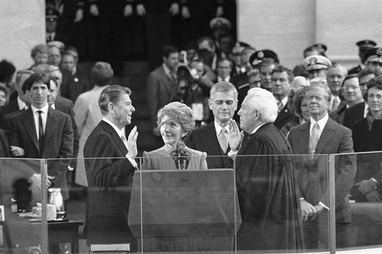 A man in a suit, in front of a crowd, raises his right hand and appears to be speaking.