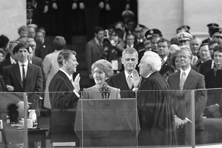 A man in a suit, in front of a crowd, raises his right hand and appears to be speaking.