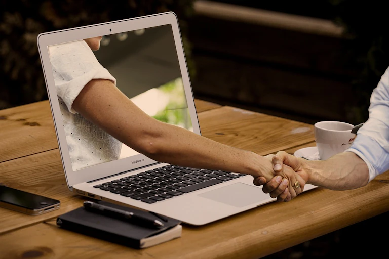 A person's arm is reaching out of the screen of a laptop on a desk to shake the hand of a person at the desk.