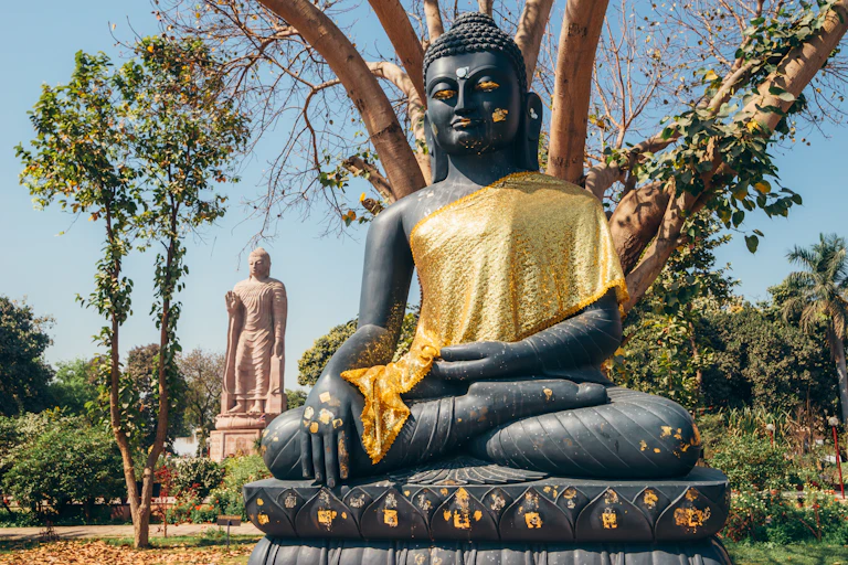 A seated and a standing Buddha statue in a garden with many trees.