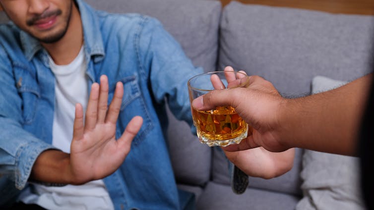 Man in a jean jacket holds his hand up to refuse a glass of alcohol