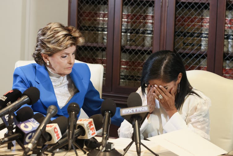 An older white woman in a blue blazer leans toward a woman with dark hair who is covering her face with her hands and sitting on front of a group of microphones.