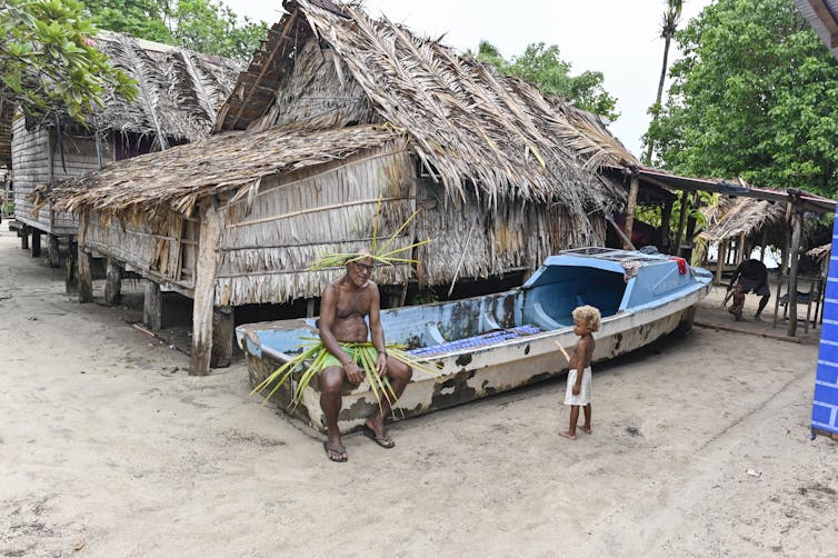 A traditional elevated house in the Solomon Islands, with an elder and a child in the foreground.