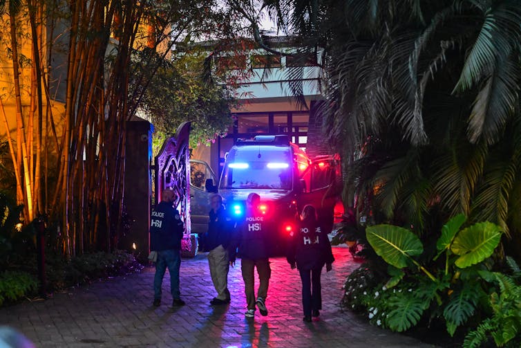 People with dark jackets are seen in front of bright blue and red lights coming from an official looking car outside of a house. Palm trees and large green bushes surrounded them.