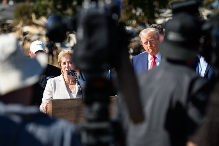 A woman is at a podium and a man is at her left nearby.