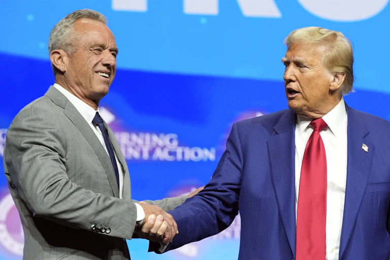 A man in a light gray suit shakes hands with a man in a navy suit and red tie