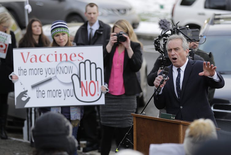 A man speaking outdoors at a podium near a woman with a sign reading: Vaccines: The more you know, the more you 'No!'