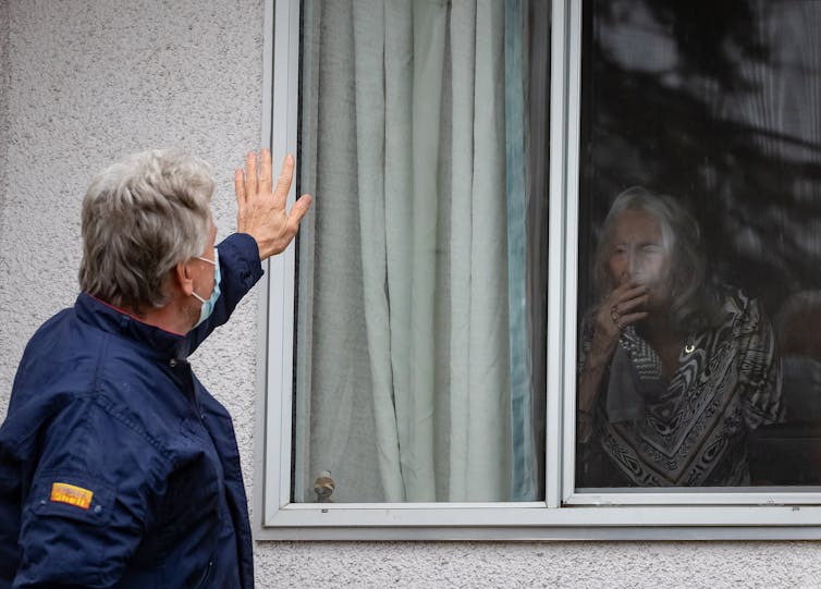 A man standing outside a window waving to a gray-haired woman inside