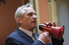A man in a suit with gray hair holding a red megaphone in front of his face