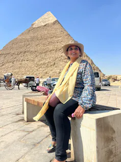 A woman sits on a bench in a hat and scarf, smiling. Behind her is a pyramid.
