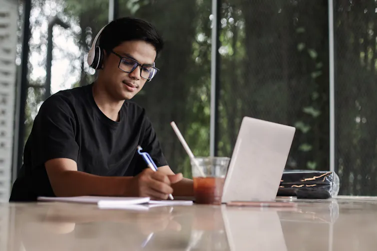 A young man wear earphones and writes, while he works with a laptop and drink.