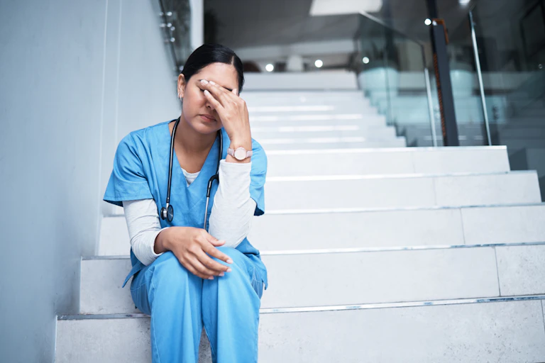 Nurse in scrubs sits on hospital stairs looking exhausted