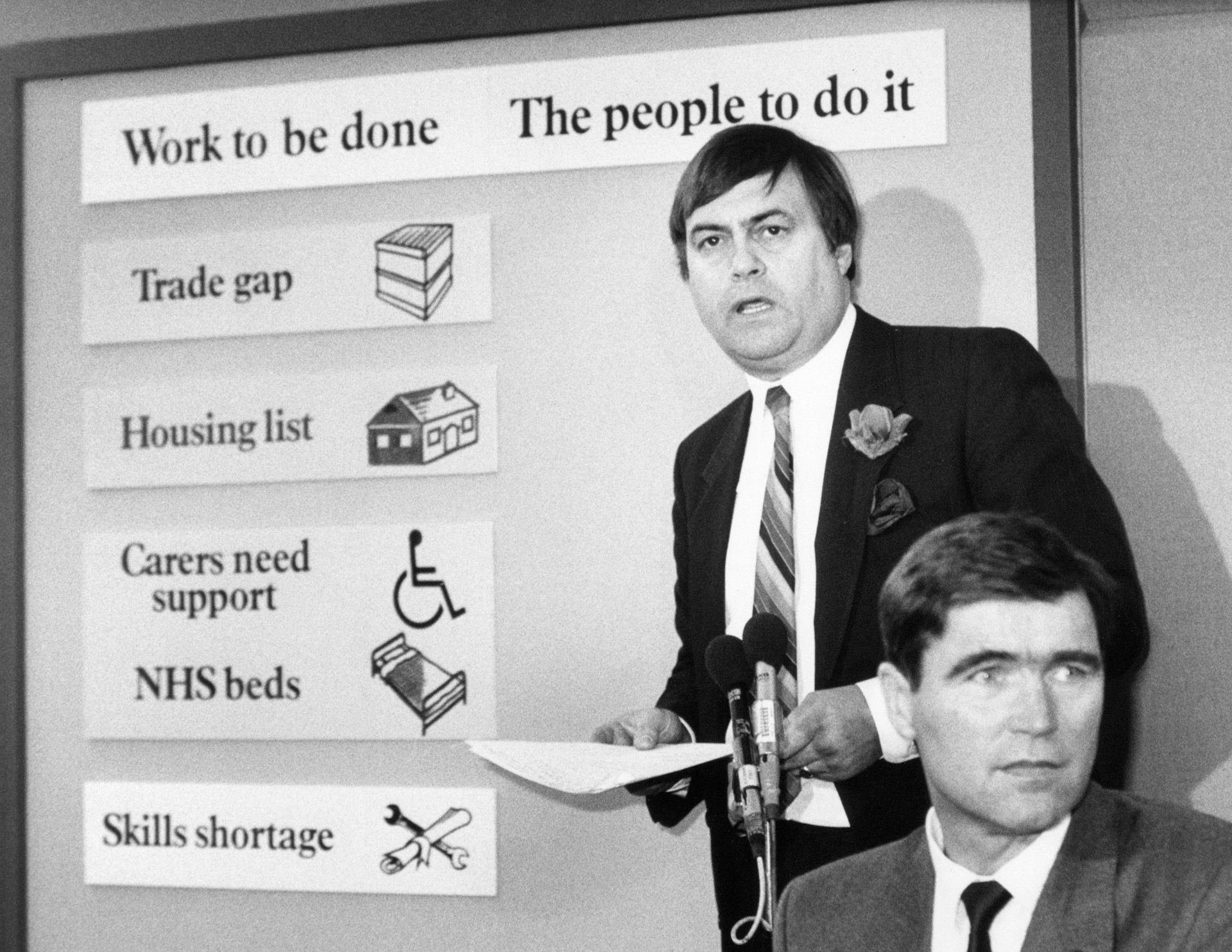 John Prescott parado entre um microfone e um quadro que diz 'Trabalho a ser feito. O povo para fazer isso'.