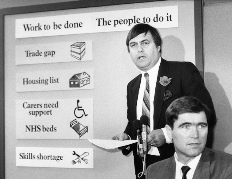 John Prescott parado entre um microfone e um quadro que diz 'Trabalho a ser feito. O povo para fazer isso'.