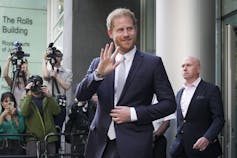 A tall slim red-haired man with a beard dressed in a suit and tie waves as he leaves a courthouse.