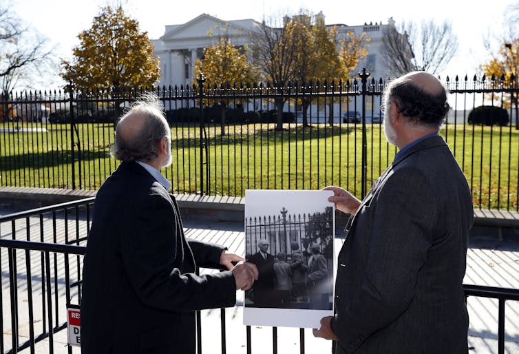 Two men in suits stand in front of the White House with their backs facing the camera, holding a black-and-white photo of two boys doing the same thing
